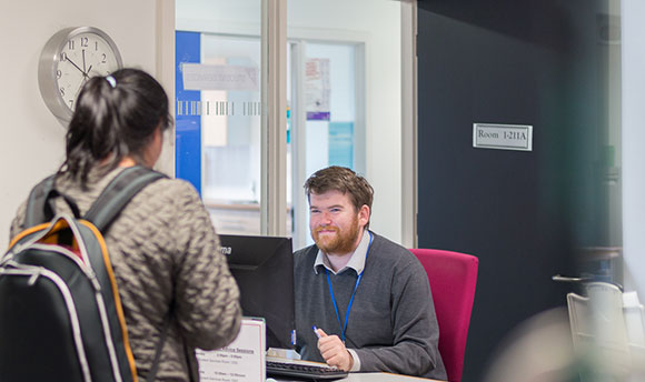A QMU staff member smiles up at a student who is in front of his desk