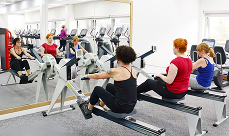 A row of women using rowing machines in front of a mirrored wall, QMU