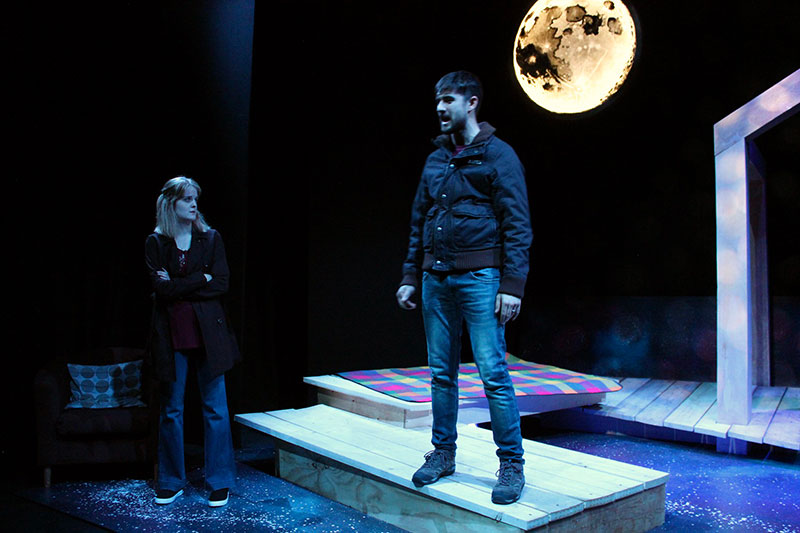 QMU drama student standing on a dark stage with a bright moon backdrop