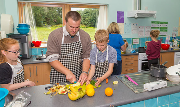 A man and some children in a kitchen wearing aprons and chopping fruit