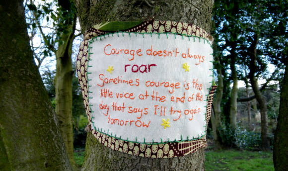 A cross stitch stuck to a tree that reads a quote about courage coming in many forms