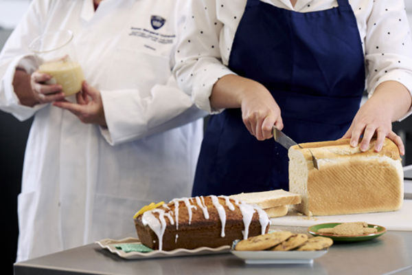 A person slicing a loaf of bread; on the counter beside them is an assortment of baked goods