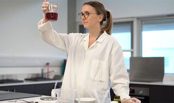 A woman wearing a lab coat, holding up a beaker of liquid.