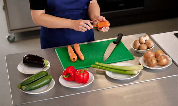 A person peels a carrot over a food preparation table laid with various vegetables.