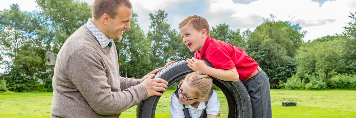 A small boy and girl playing with a tyre in a playpark a man holding the tyre so the girl can climb through the middle