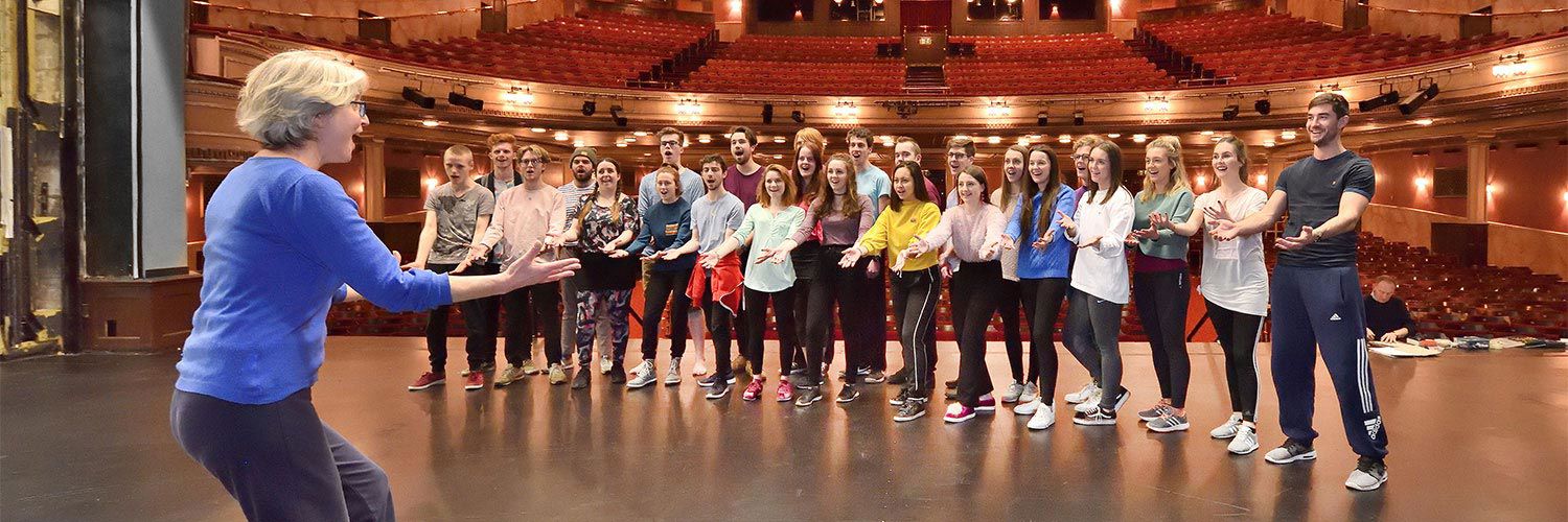 A group of students replicate a lecturer's movement of holding their hands outstretched. They are all stood on a theatre stage.