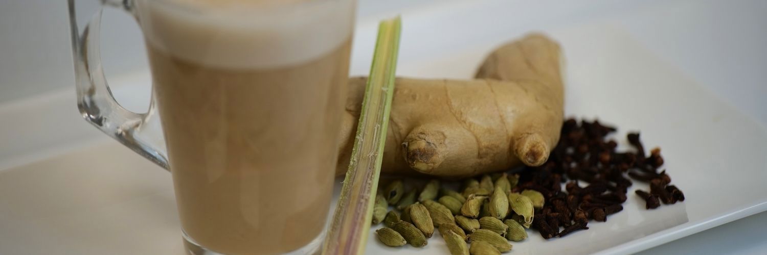Chai tea in a clear coffee glass with cut lemongrass, ginger, cardamom and tea leaves beside