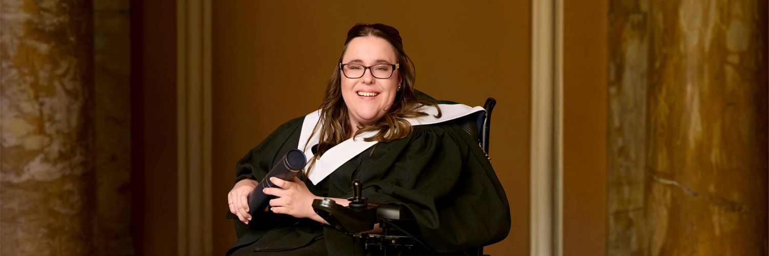portrait style image of Charlotte sitting in wheelchair  wearing graduation robe and holding graduation scroll 
