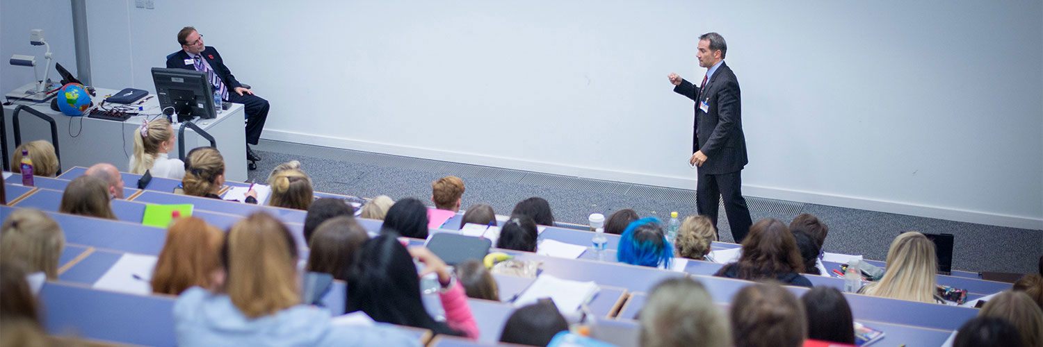 A faraway shot of a person delivering a lecture to a hall full of students, the photo is taken from the top of the lecture hall.