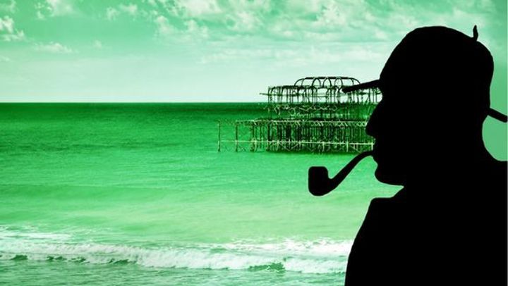 Green-toned silhouette of a detective with a pipe overlooking the remains of Brighton West Pier against a cloudy seaside sky