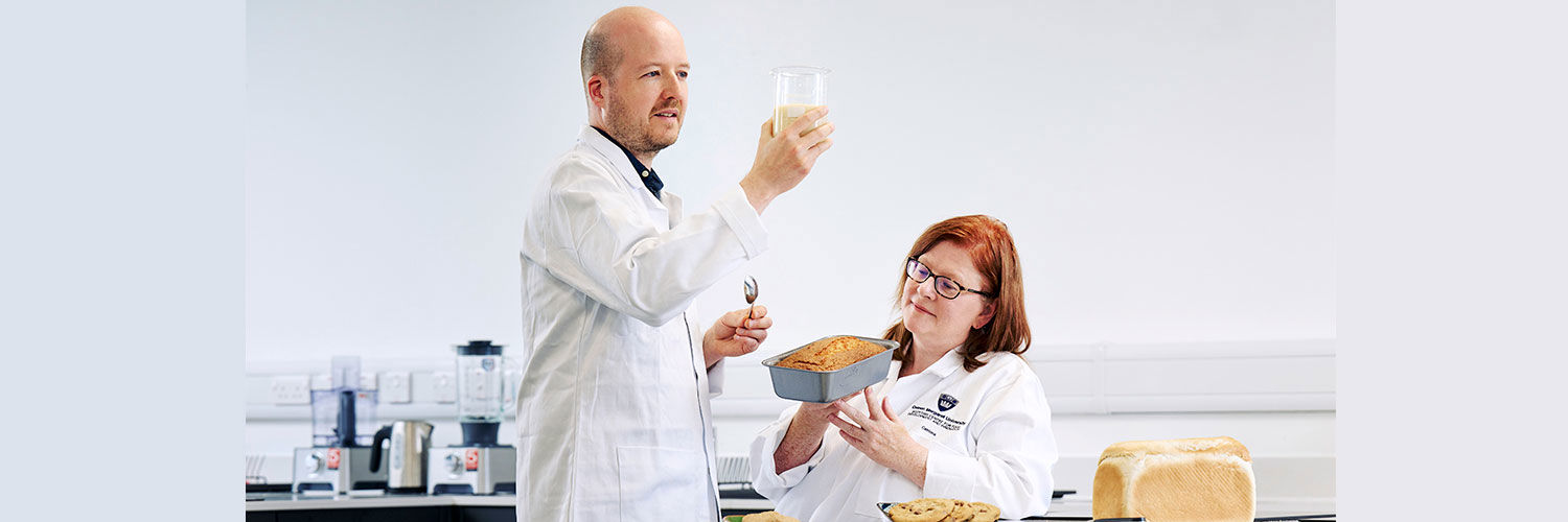 A male and female scientist dressed in white lab coats look at a creamy substance in a glass beaker and examine a cake in a loaf tin while in a test kitchen