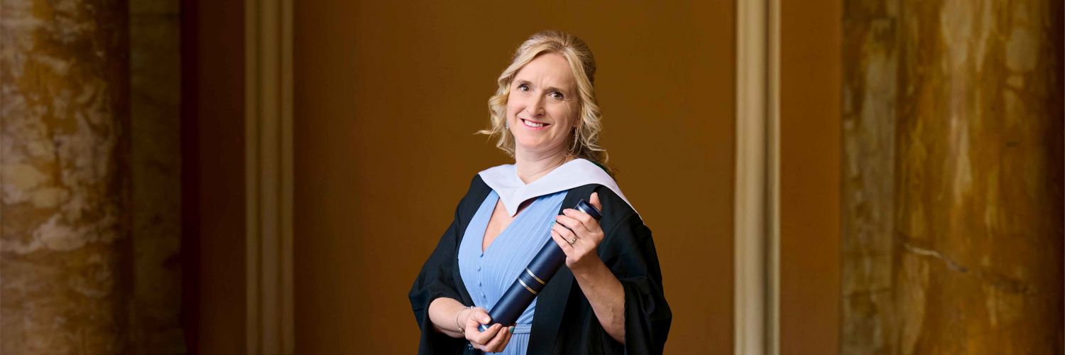 portrait style image of female blonde student wearing graduation robe and holding graduation scroll 