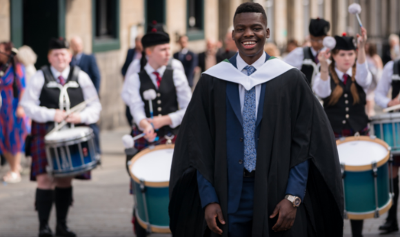 a young black man with a big smile, wearing his graduation gown and standing in front of a pipe band playing outside the graduation venue