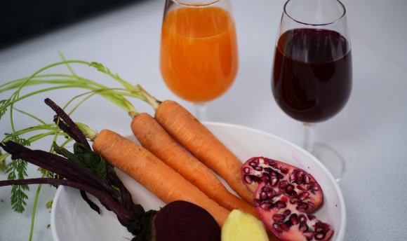 One glass with vibrant orange liquid and one glass with blackcurrant purple liquid, three carrots, one beetroot, one piece of ginger, one pomegranate 