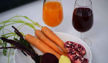 One glass with vibrant orange liquid and one glass with blackcurrant purple liquid, three carrots, one beetroot, one piece of ginger, one pomegranate 