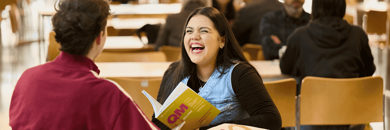 Male and female student laughing in the canteen holding a QMU prospectus. 