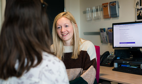 Two women talking by a computer desk, the woman in view is smiling kindly at the other