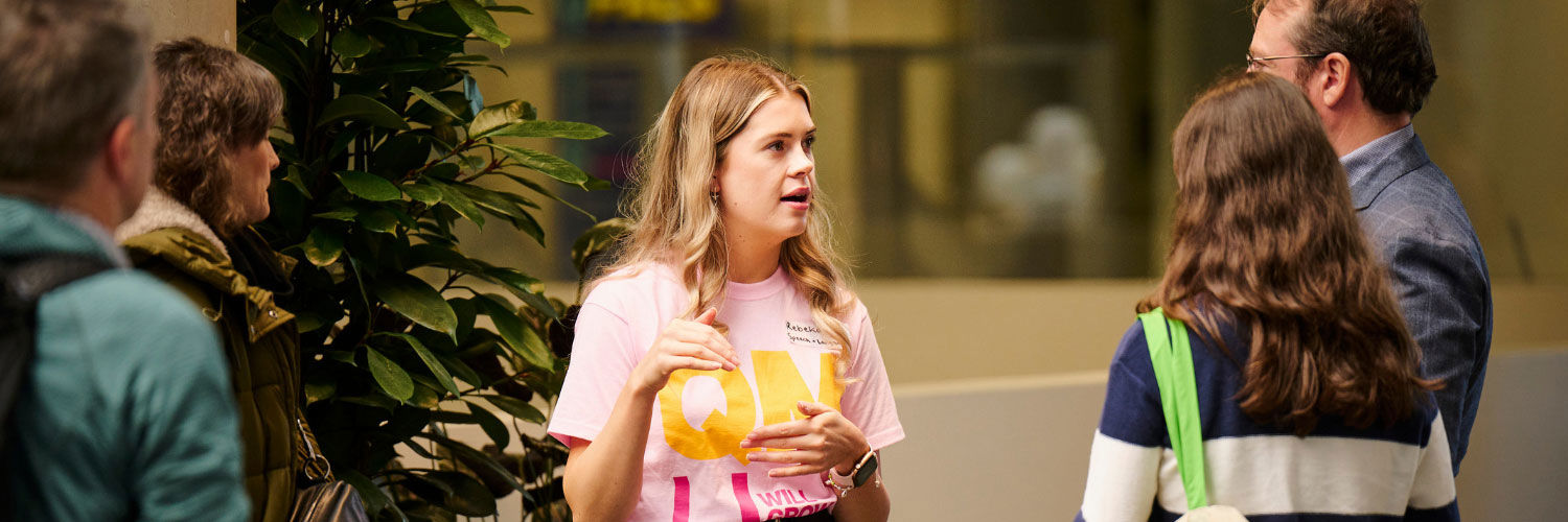 A young woman wearing a QMU t-shirt speaks to prospective students on a campus tour.