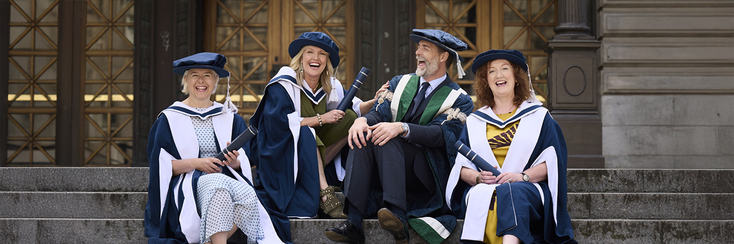 Four people in graduation gowns and hats smiling whilst sat on stone steps