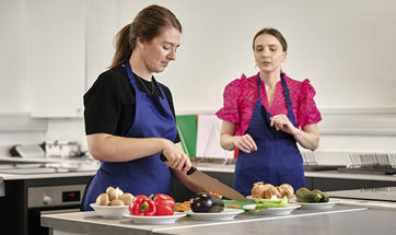 Two females chatting whilst ones cuts up vegetables