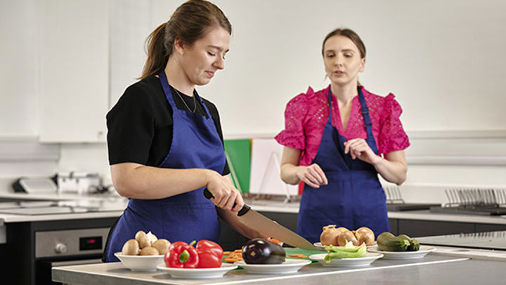 Two females chatting whilst ones cuts up vegetables