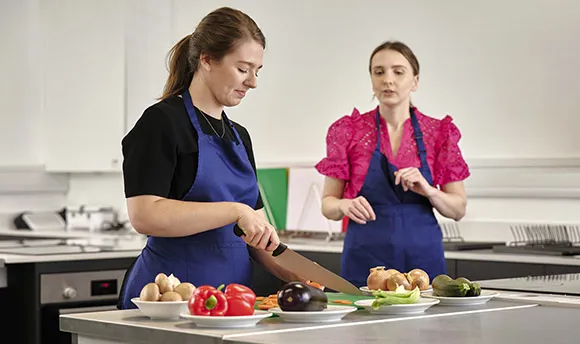 Two females chatting whilst ones cuts up vegetables