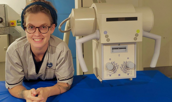 image of a young woman in grey health professionals uniform smiling next to a radiography machine