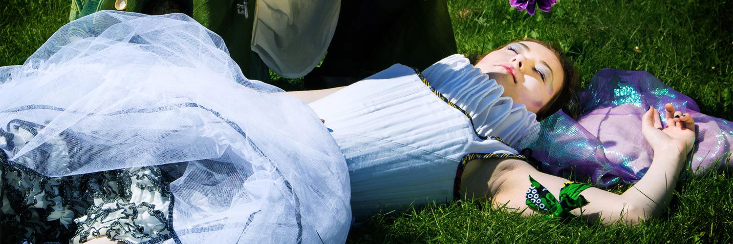 Two actors wearing period-appropriate Shakespearian outfits for an outdoor performance of A Midsummer Night's Dream. The woman lays sleeping while the male actor hovers beside her.