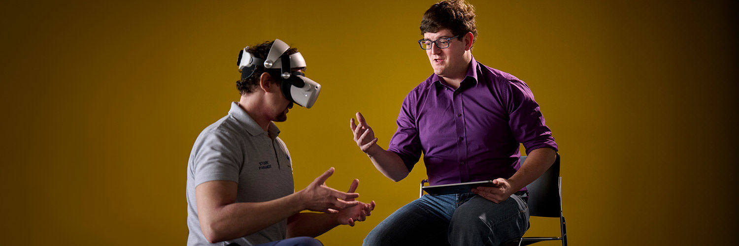 A man is seated, demonstrating the movement required in a VR paramedic simulation. They are sat alongside another man who is wearing a VR headset.