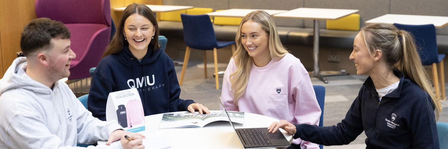 Four students wearing branded QMU merchandise sit at a table enjoying a chat together.