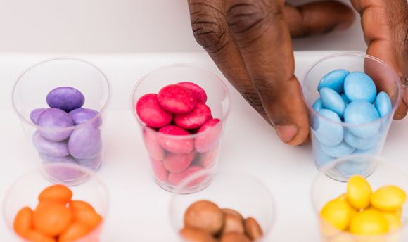 A human hand reaches towards one of 10 tiny containers full of brightly coloured samples of sweets. Each container contains individual colours of sweets.
