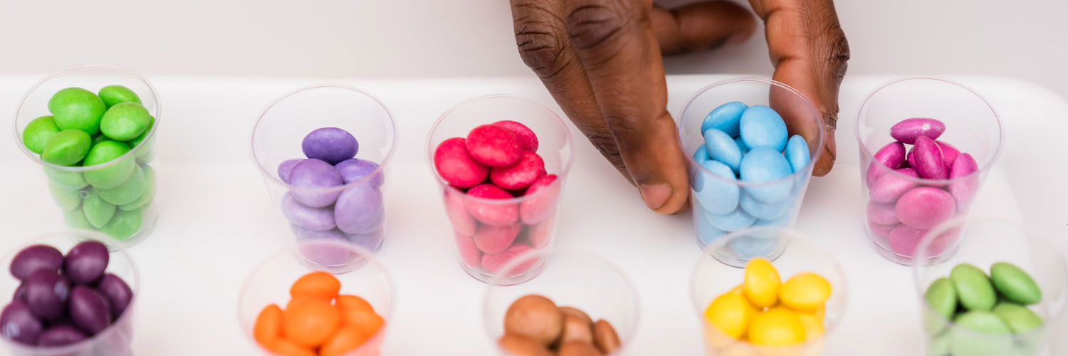 A human hand reaches towards one of 10 tiny containers full of brightly coloured samples of sweets. Each container contains individual colours of sweets.