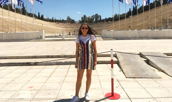 A person poses for a photo, various Greek and Olympics flags stand on tall flagpoles behind them.