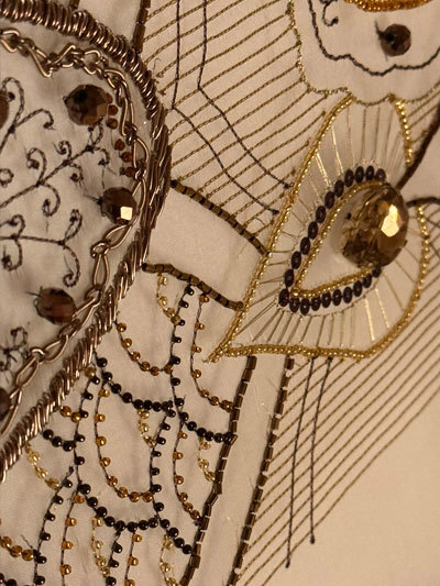 A close up of a hand-embroidered piece of fabric. The item is cream in colour, while the beads are a variety of shades including gold, brown, and black.