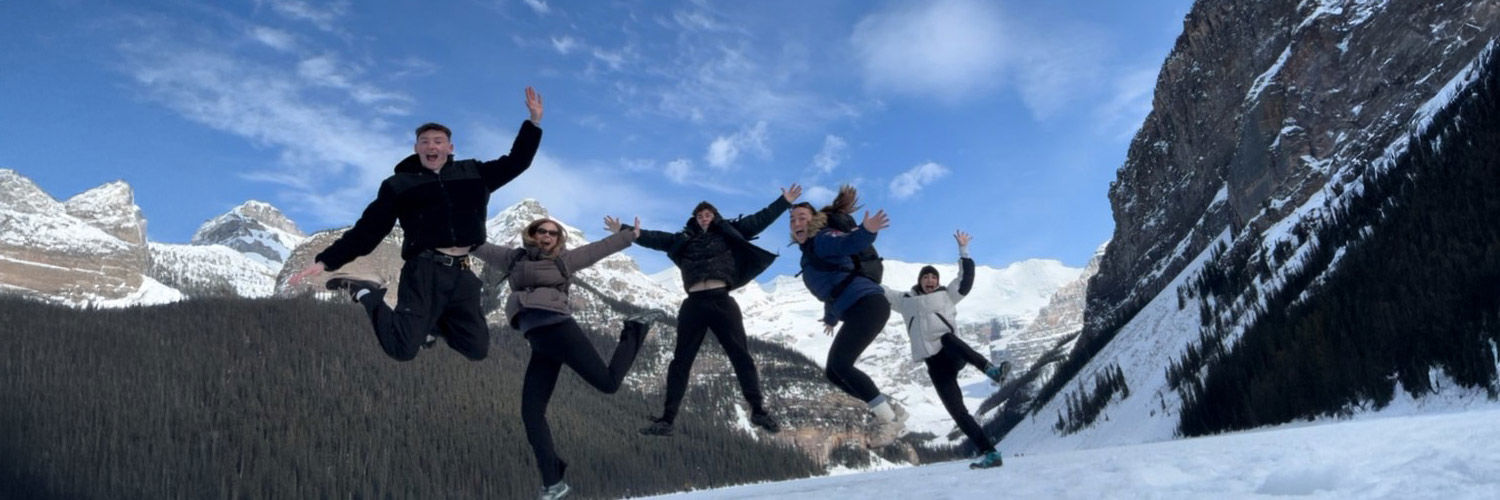 A group of people mid-jump, there is a vast snowy mountain range behind them.