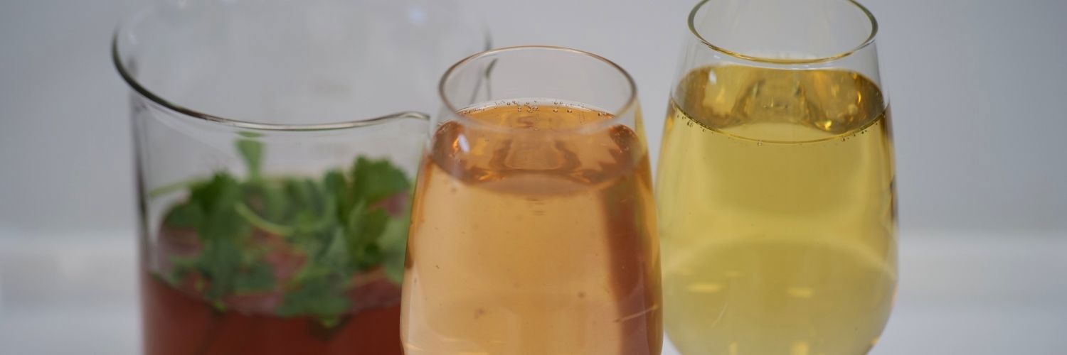 One glass with peach liquid, one glass with yellow liquid and one beaker behind with dark red liquid with coriander leave on top