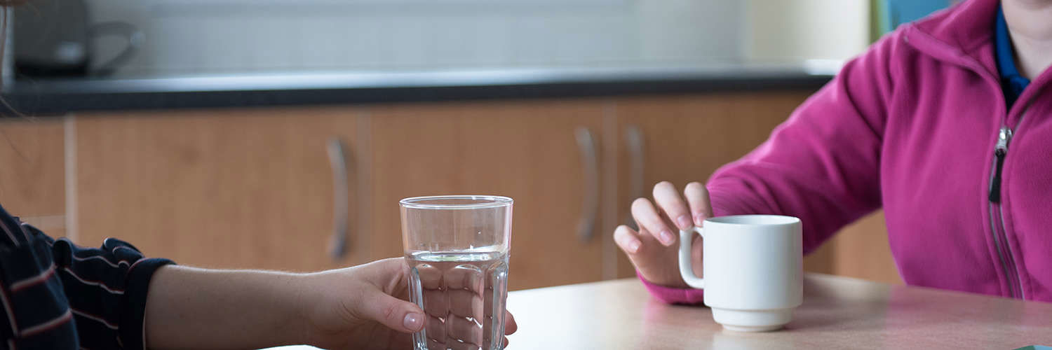 Close up of two people sat at a table, one has a glass of water and the other a mug