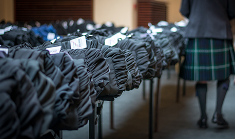 Rows of graduation gowns folded on tables, a man in a kilt stands to the side
