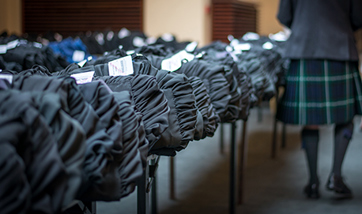 Rows of graduation gowns folded on tables, a man in a kilt stands to the side