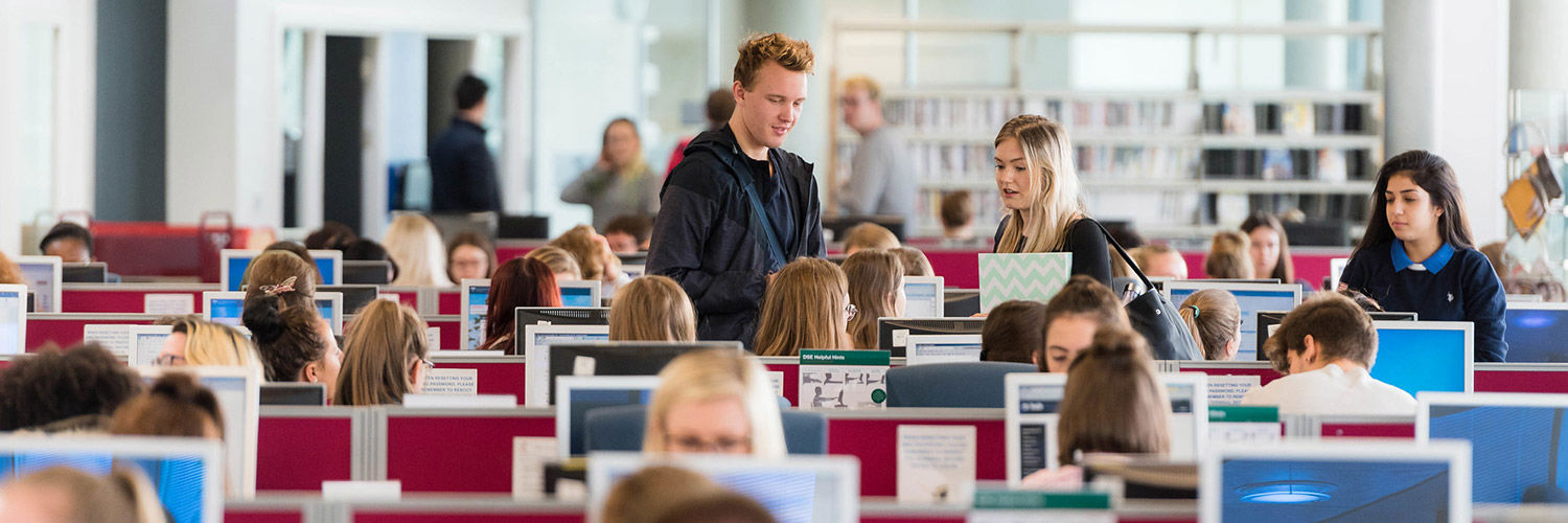 Two QMU students standing talking to another by their computer desk in a busy room