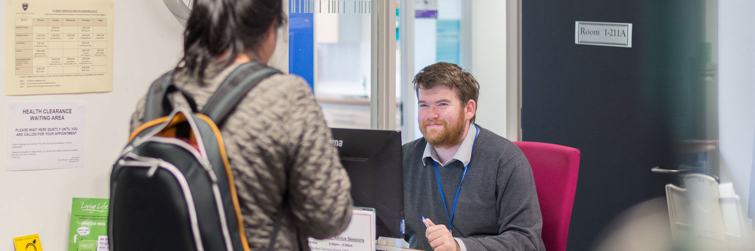 A QMU staff member smiles up at a student who is in front of his desk