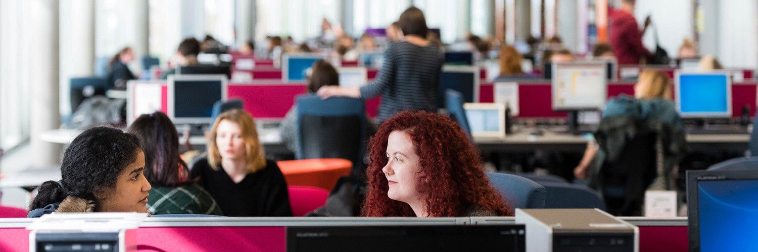 Two QMU students talking whilst working on their desktop computers in a busy classroom