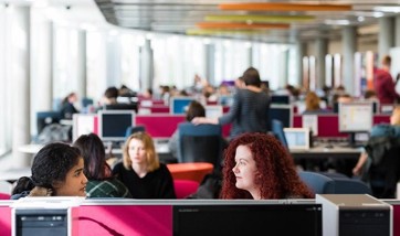 Two QMU students talking whilst working on their desktop computers in a busy classroom