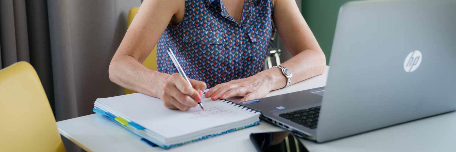 A woman sitting over a laptop and writing in a notebook
