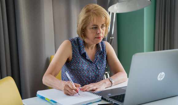 A woman sitting over a laptop and writing in a notebook