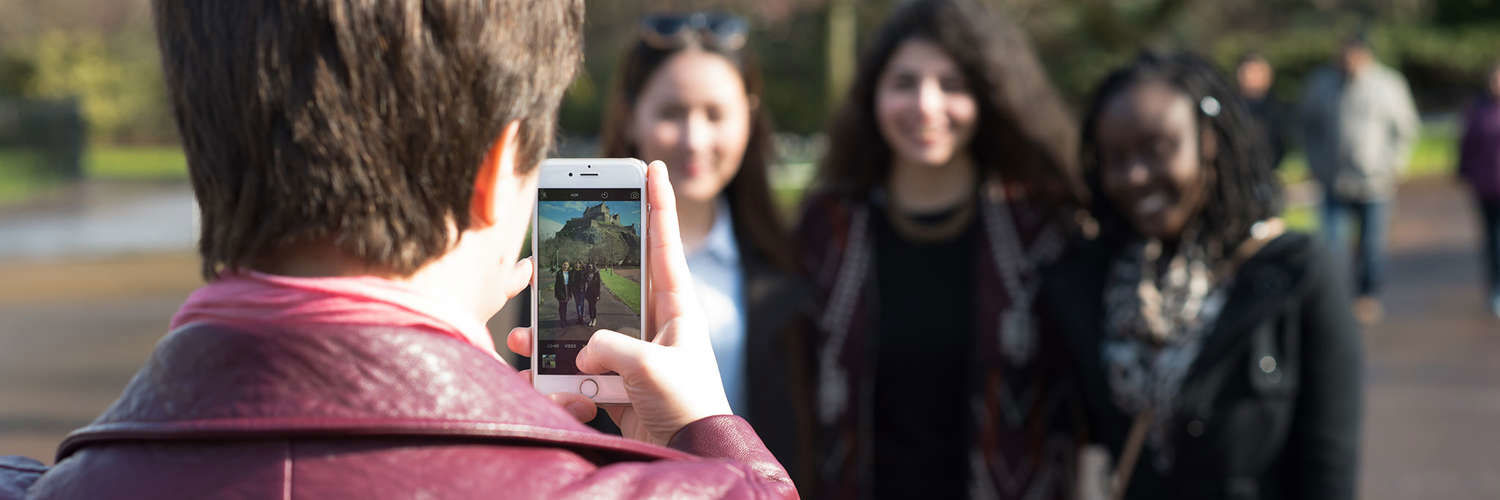 A photo is being taken of 3 girls, on the phone screen Edinburgh Castle is in the distance
