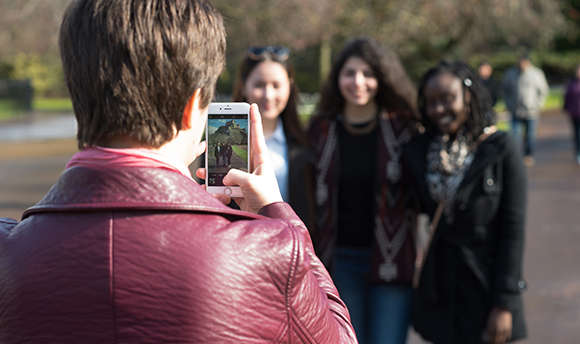 A photo is being taken of 3 girls, on the phone screen Edinburgh Castle is in the distance