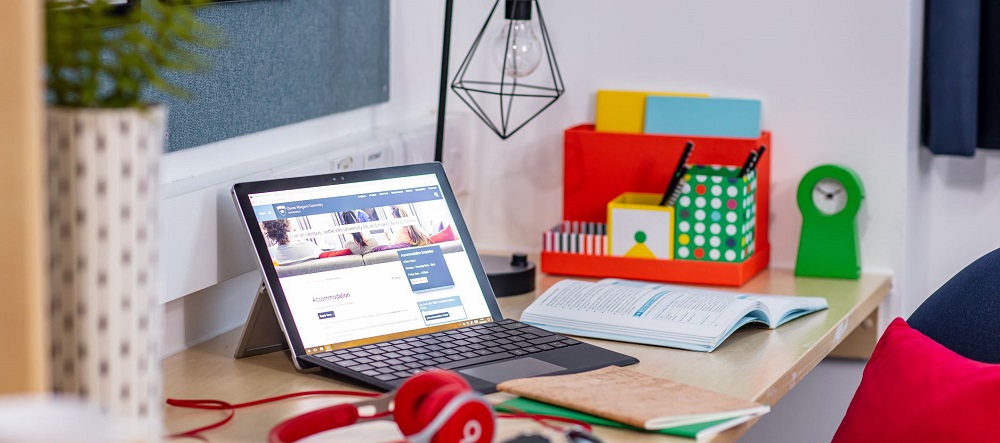 A laptop on someone's desk at home beside a set of headphones and some notepads