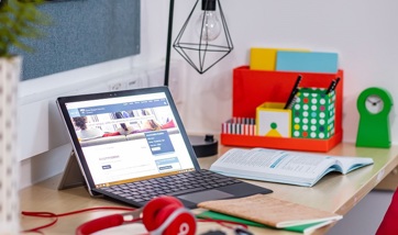 A laptop on someone's desk at home beside a set of headphones and some notepads