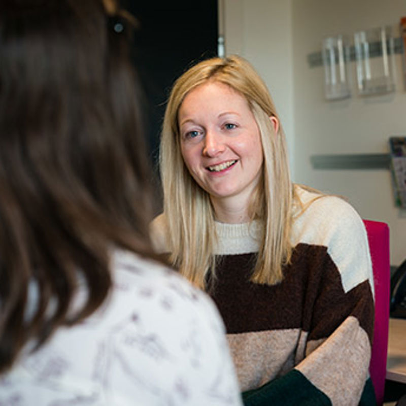 Two women talking by a computer desk, the woman in view is smiling kindly at the other